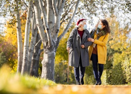An elderly woman walks with a cane in one hand and her other arm linked around her caregiver's arm