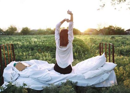 A young woman stretches while sitting on her bed in the middle of an open field.