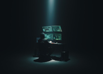 A person wearing a hoodie sits in front four computer monitors under a spotlight
