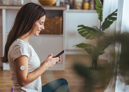 A woman with a continuous glucose monitor on her arm sits on the floor looking at her phone while smiling