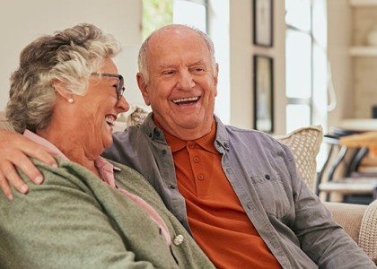 An elderly couple sit on the couch laughing