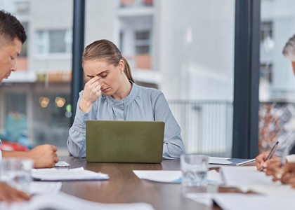 A woman sits at the head of a conference table pinching her nose with her laptop open